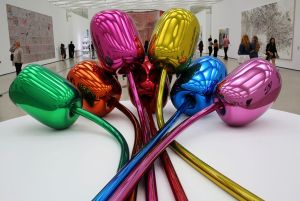 An installation view shows a shiny, brightly colored sculpture of oversized metallic balloon-like flowers displayed in a white museum gallery.