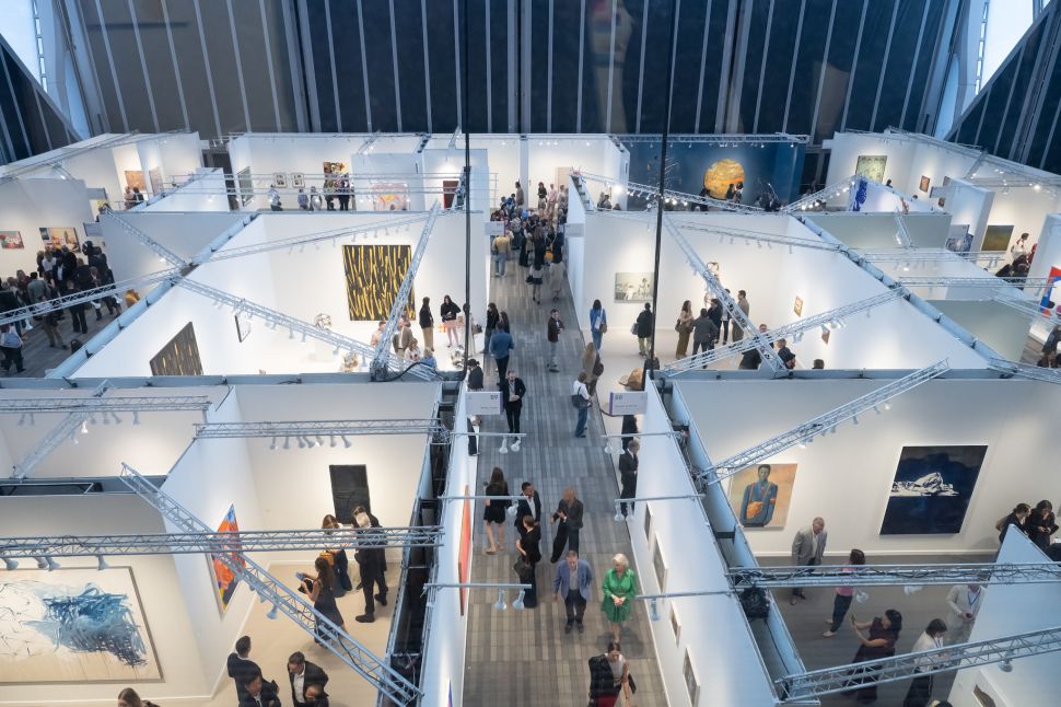 Overhead view of a bustling contemporary art fair inside a large, glass-walled venue, showing rows of white-walled booths filled with paintings, sculptures, and visitors engaging with the artworks and gallerists.
