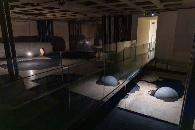 An installation view of Alicja Kwade’s "INFRASUPRA" shows a dim underground space at FOROF with a suspended stone hovering above a dark circular form on the floor, illuminated by a small bright light.