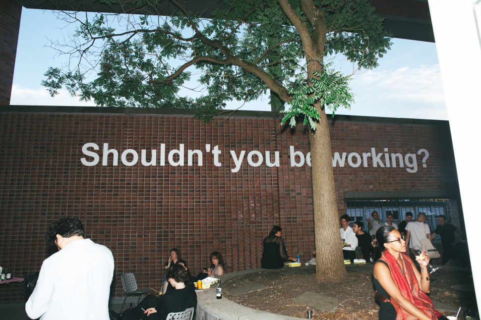 Visitors gather in a courtyard beneath a large wall text reading “Shouldn’t you be working?” on a brick facade.