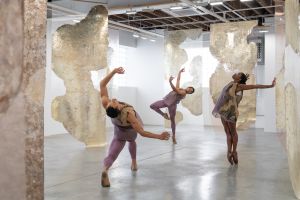 A performance of Echoes on the Wall shows three dancers in purple costumes moving through Rosalind Tallmadge’s sculptural installation of translucent panels in a white-walled gallery space.
