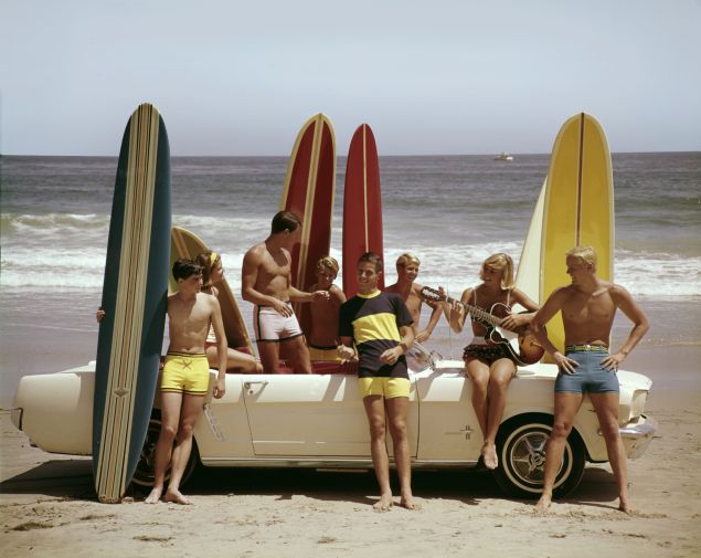 vintage photo of several men and women in swimwear with surfboards standing in front of ford mustang on the beach