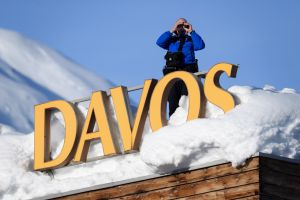 A uniformed security officer stands on a snow-covered rooftop above a large yellow DAVOS sign, scanning the alpine landscape through binoculars against a clear blue sky.