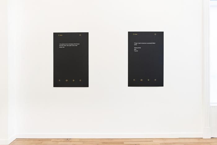 An installation view shows two black rectangular text-based works mounted on a white gallery wall above a wooden floor.