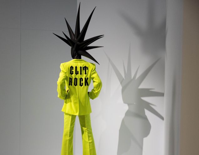 A museum visitor stands beside a mannequin wearing a neon yellow suit with “Clit Rock” printed on the back and a spiky black headpiece casting a shadow on the wall.