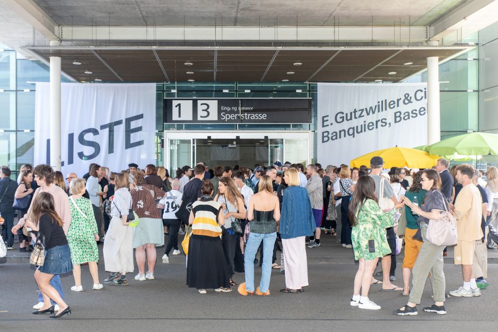 A large crowd gathers outside the Liste entrance in Basel, with white fair banners hanging on either side of the building and colorful umbrellas to the right.