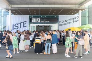 A large crowd gathers outside the Liste entrance in Basel, with white fair banners hanging on either side of the building and colorful umbrellas to the right.