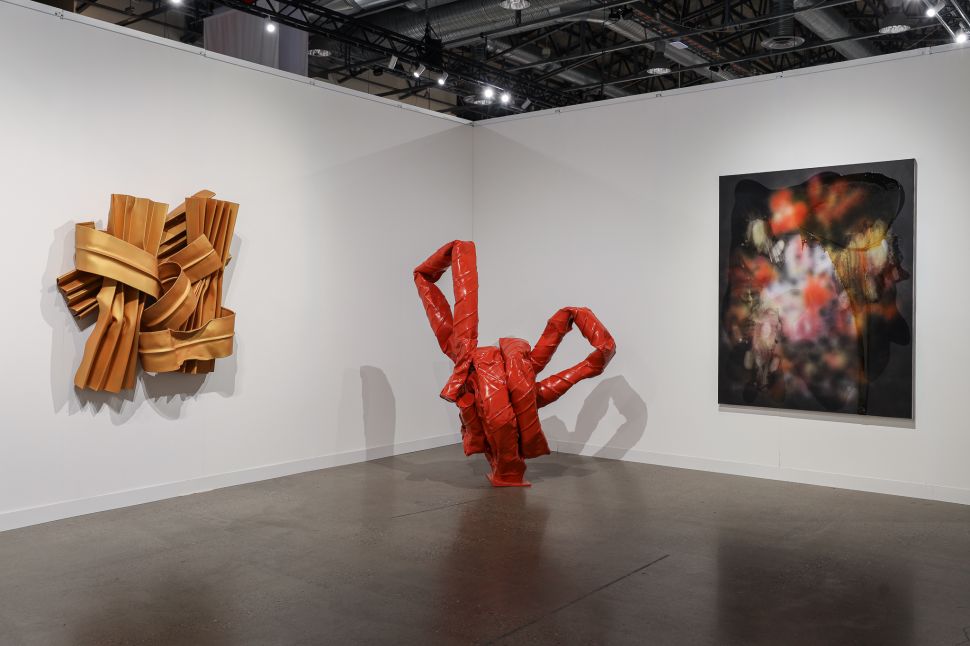 A gallery booth presents three large-scale contemporary works, including a gold wall sculpture, a red twisting sculpture at center and an abstract painting with blurred red and black forms.
