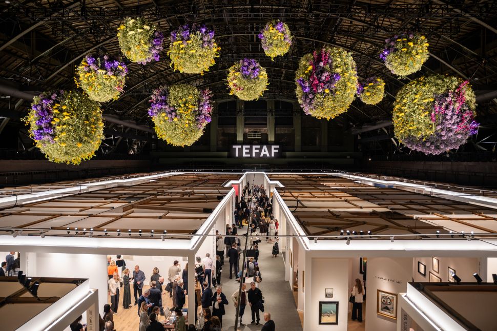 An overhead view of TEFAF New York shows visitors moving through the fair’s aisles beneath large suspended floral installations and an illuminated TEFAF sign.