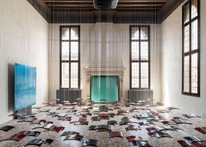 A spacious gallery installation with suspended vertical wires, colorful curved glass panels arranged across the floor, and large wall-mounted glass works.