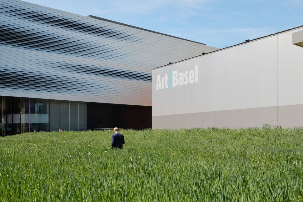 A man in a suit walks through a tall grassy field outside Art Basel’s contemporary Messeplatz venue, with a sleek, patterned metal building in the background.
