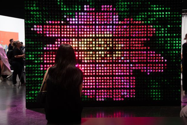 A visitor sits in front of Hito Steyerl’s Green Screen, a glowing LED wall of red, pink and green lights.