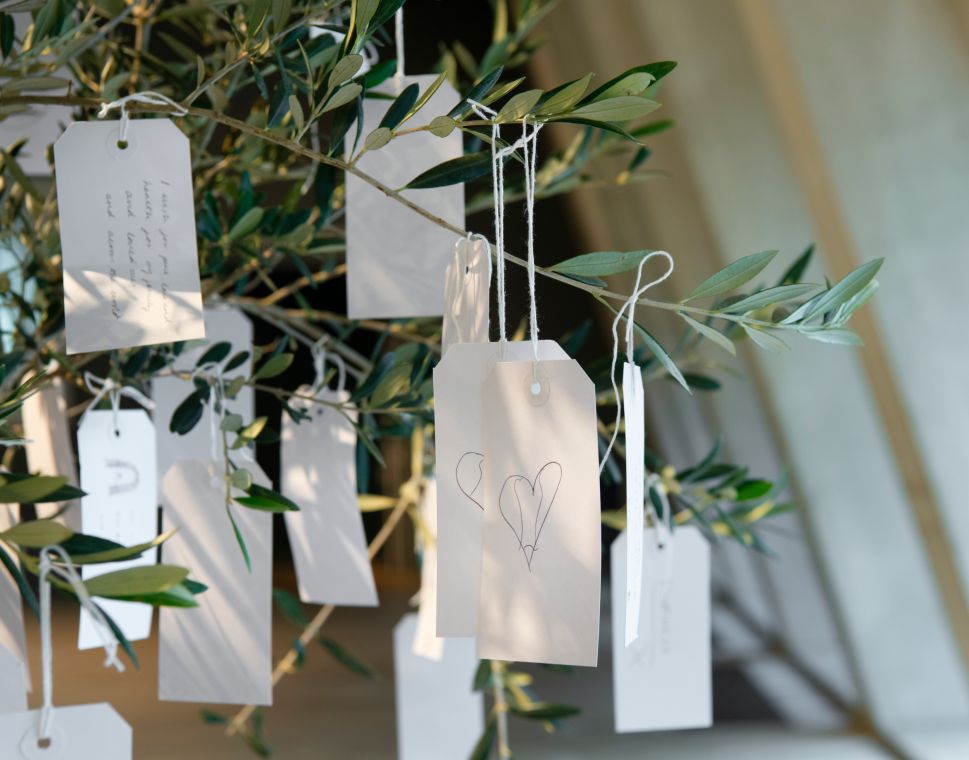 A close-up photograph shows handwritten white wish tags tied with string to leafy branches, including one tag with a simple drawing of two hearts.