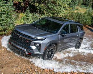 A gray Chevrolet Traverse SUV driving through a shallow creek, splashing water around the wheels, surrounded by forested terrain.